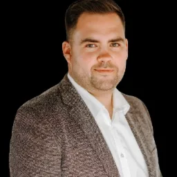 Portret mężczyzny w tweedowej marynarce i białej koszuli na czarnym tle, z lekkim uśmiechem i krótkimi, schludnie ułożonymi włosami.