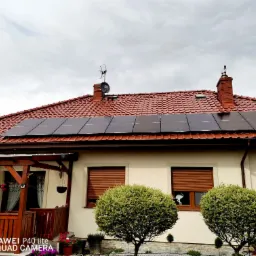 Dom jednorodzinny z zainstalowanymi panelami słonecznymi na czerwonym dachu, widok z przodu, z dekoracyjnymi krzewami i drewnianą pergolą.