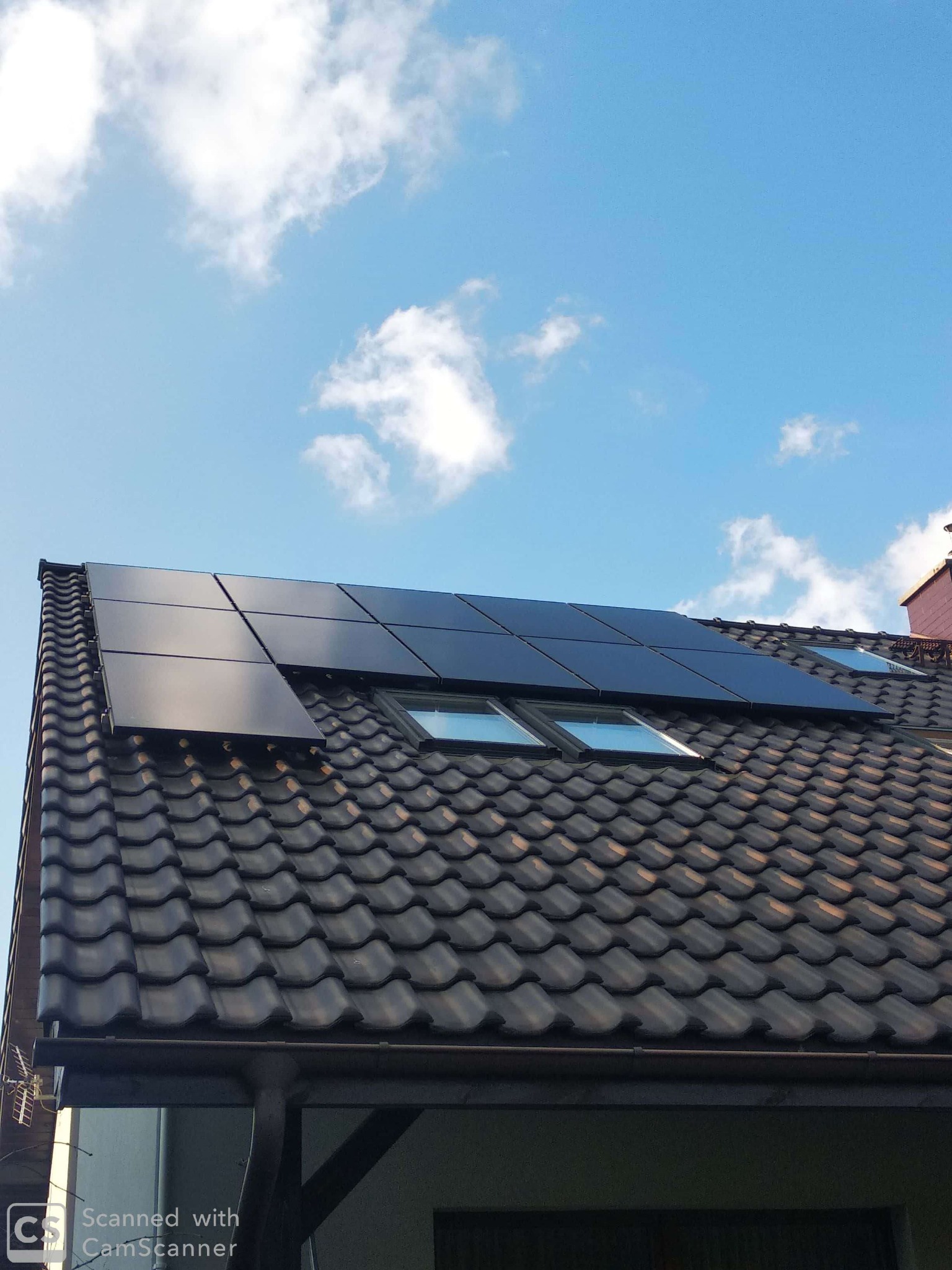Czarne panele fotowoltaiczne zamontowane na ciemnym, skośnym dachu z dachówką ceramiczną, widoczne okna dachowe, błękitne niebo z białymi chmurami w tle, antena telewizyjna po lewej.