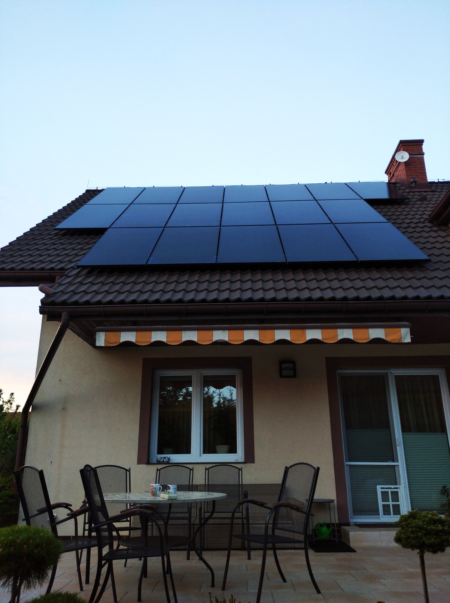 Panele słoneczne na brązowym dachu domu jednorodzinnego z tarasem, widoczne krzesła i stół na zewnątrz.