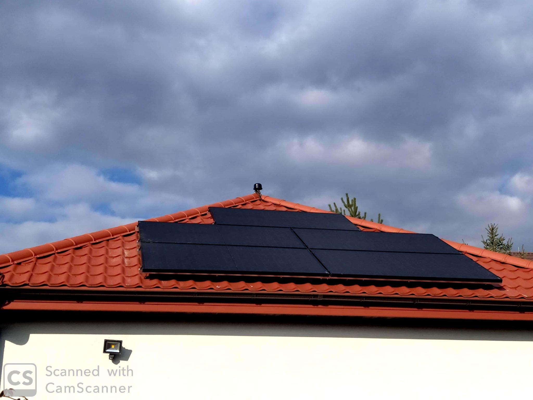 Część dachu pokryta czerwoną dachówką z zainstalowanymi panelami słonecznymi w układzie prostokątnym, na tle pochmurnego nieba.
