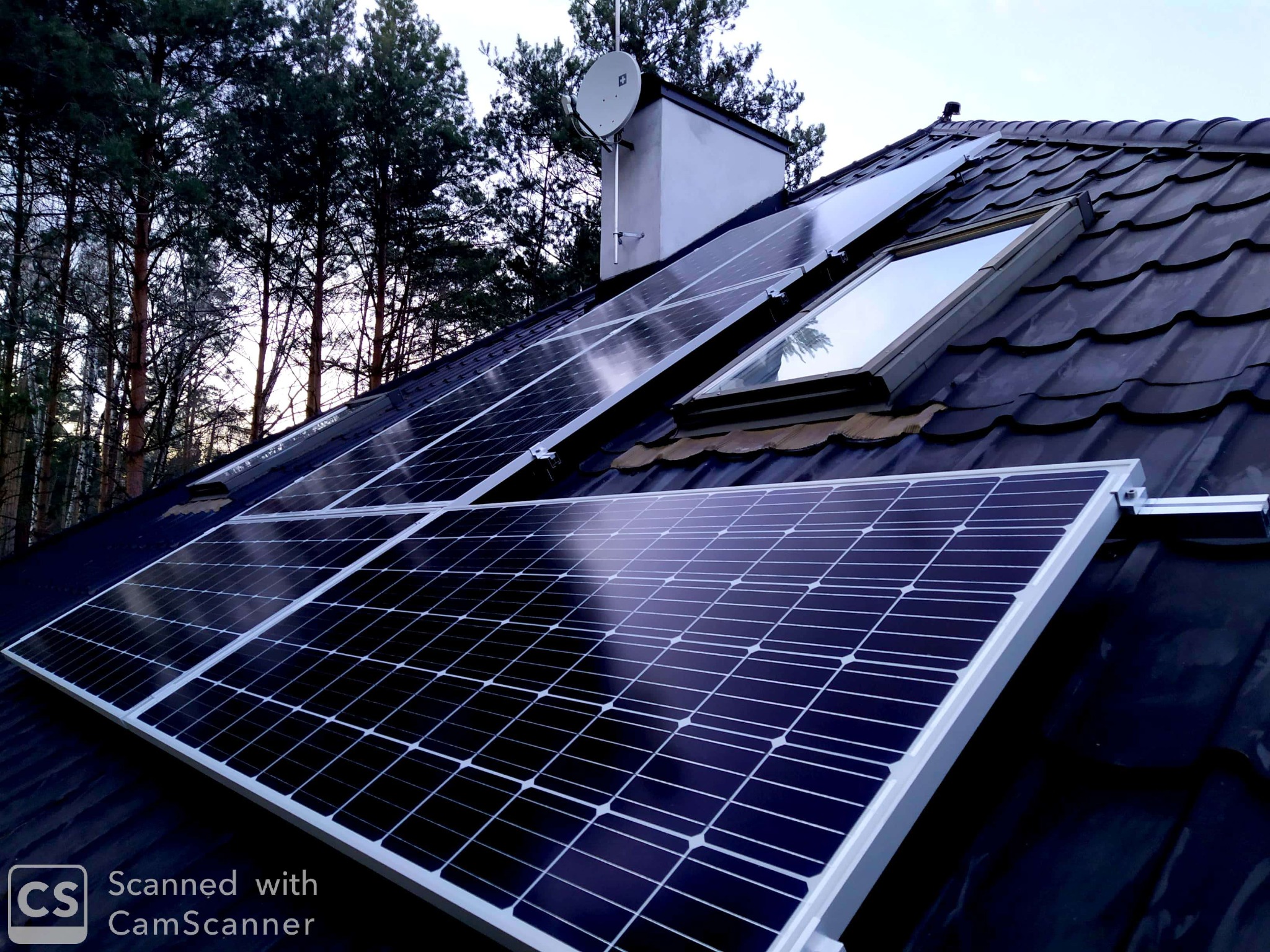 Panele słoneczne zamontowane na ciemnym dachu z dachówki ceramicznej, obok okna dachowego i komina z anteną satelitarną, w tle drzewa.