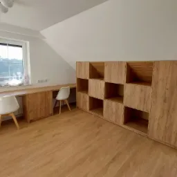 Szafa wnękowa z otwartymi półkami i biurko na wymiar z blatem z drewna w odcieniach brązu, dopasowane do skośnego sufitu na poddaszu, uzupełnione białymi krzesłami.