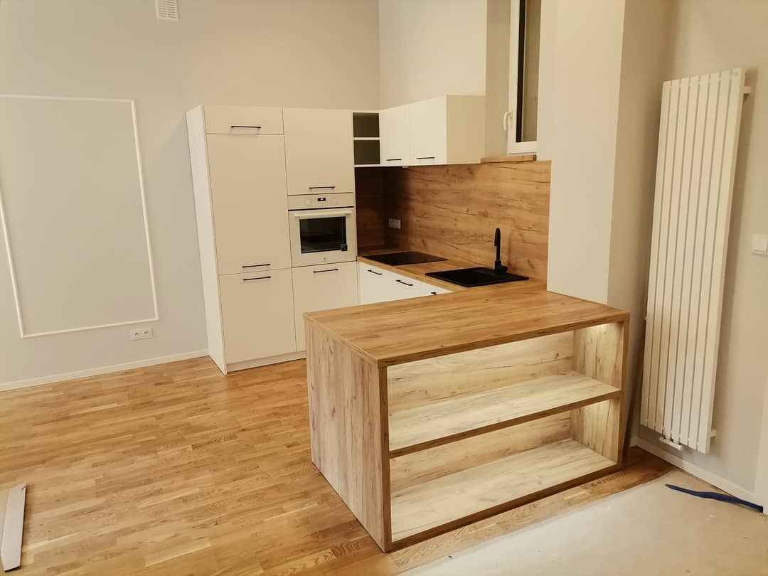 Nowoczesna kuchnia z białymi szafkami, drewnianym blatem i wyspą z podświetlanymi półkami, na parkiecie. Widoczny fragment grzejnika.