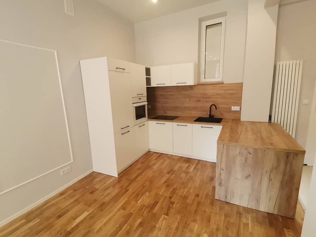 Nowoczesna kuchnia z białymi szafkami, drewnianym blatem i podłogą, wysoka zabudowa z piekarnikiem, czarny zlew i bateria, minimalistyczny styl.