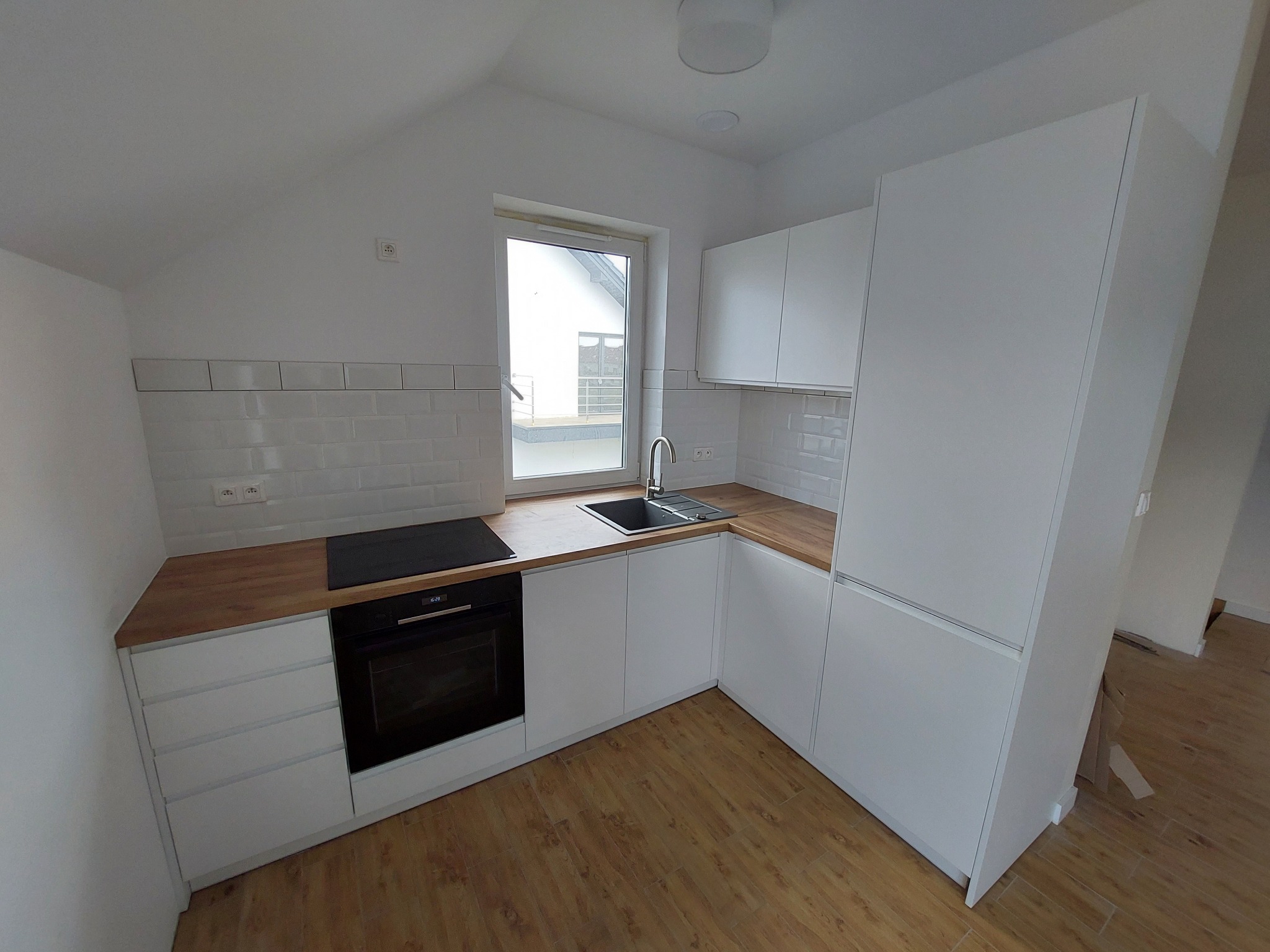 Nowoczesna, biała zabudowa kuchenna z drewnianym blatem, płyta indukcyjna i piekarnik w aneksie kuchennym pod skośnym sufitem z oknem.