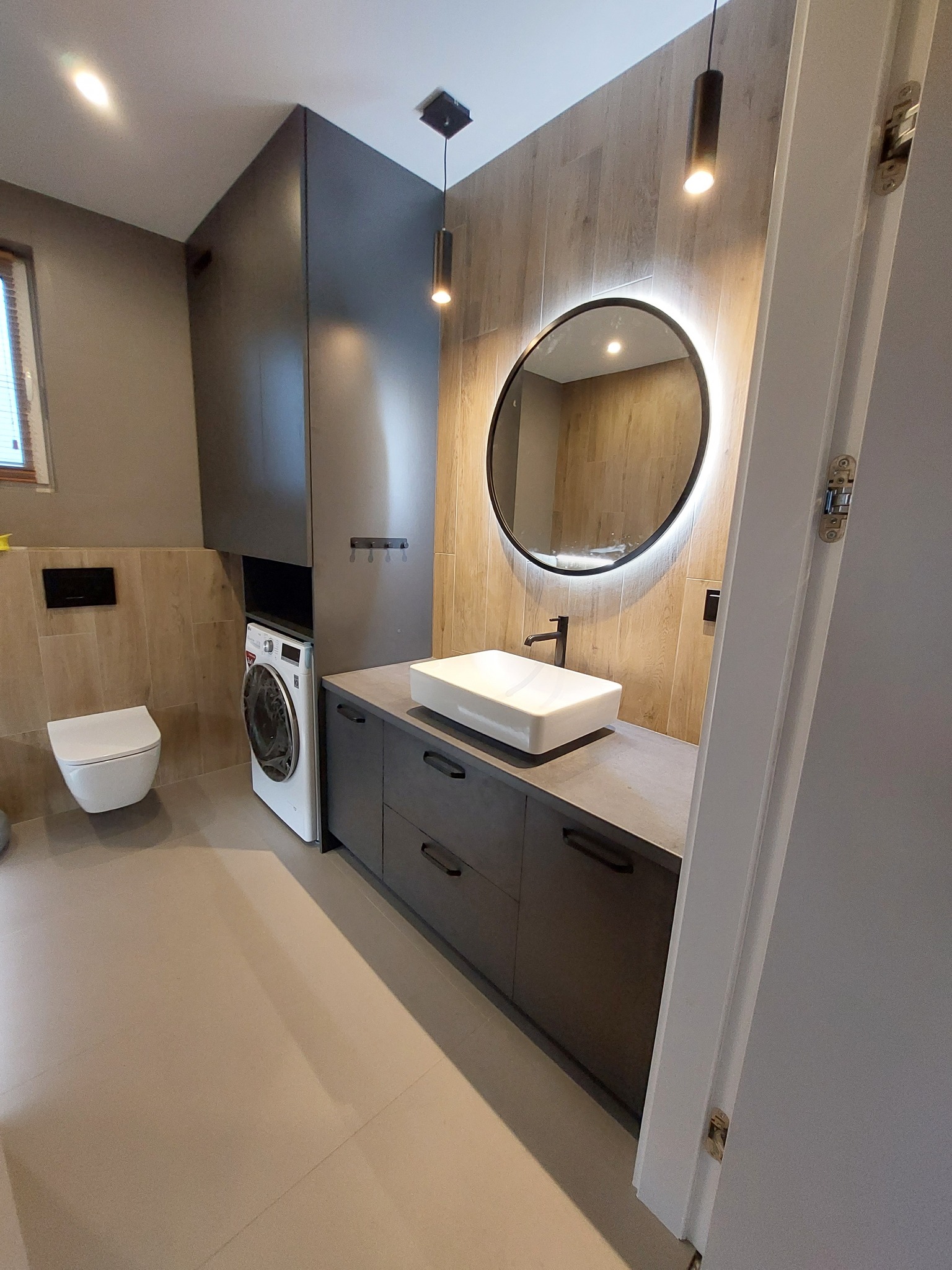 Nowoczesna łazienka z zabudową meblową w odcieniach szarości, z pralką wkomponowaną w szafę, okrągłe lustro z podświetleniem LED, umywalka nablatowa i wisząca toaleta.