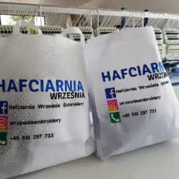 Dwie białe torby z nadrukiem 'Hafciarnia Września' oraz ikonami mediów społecznościowych i numerem telefonu, na tle maszyny do haftowania.