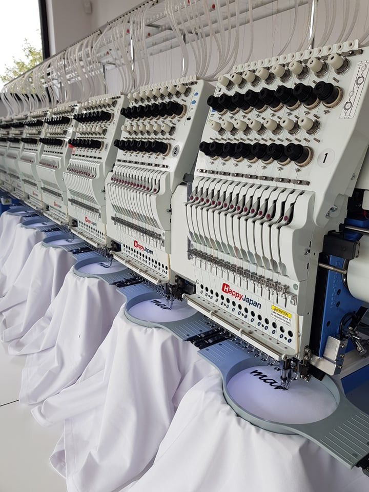 Wielogłowicowa maszyna hafciarska Happy Japan w trakcie pracy, z bliska widoczne białe koszulki z czarnym haftem. W tle widoczne rzędy szpul z nićmi.