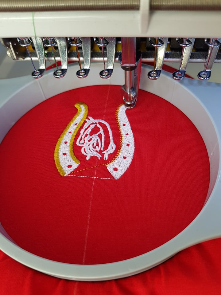 Haftowanie logo na czerwonym materiale za pomocą automatycznej hafciarki. Widoczna igła w trakcie pracy i fragmenty wielokolorowych nici.