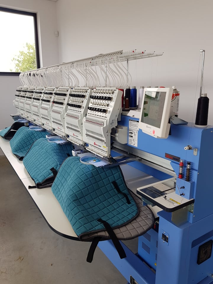 Wielogłowicowa maszyna hafciarska w trakcie pracy nad serią turkusowych, pikowanych pokrowców na siodła, widoczne nici, szpulki i ekran dotykowy maszyny.