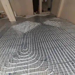 Białe rury ogrzewania podłogowego ułożone na srebrnej folii z nadrukiem KELLER, przymocowane do czarnej siatki w surowym wnętrzu budynku w trakcie budowy.