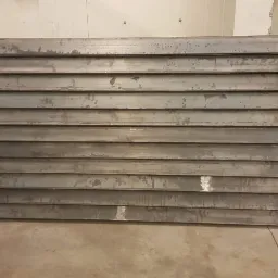 Surowa, metalowa konstrukcja z poziomych profili, prawdopodobnie fragment bramy lub ogrodzenia, w hali produkcyjnej, widoczne ślady obróbki i niedoskonałości powierzchni.