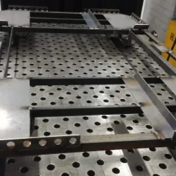 Konstrukcja metalowa spawana na perforowanym stole spawalniczym, widoczny żółty transformator spawalniczy w tle.