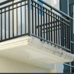 Ciemna, metalowa balustrada balkonowa z pionowymi szczeblami na tle jasnej elewacji budynku z dużymi, odbijającymi światło oknami.