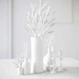 Monochromatyczna kompozycja dekoracyjna w bieli: wysokie źdźbła zboża w cylindrycznym wazonie, dyfuzor zapachowy z patyczkami, mały bukiet kwiatów, dekoracyjne jabłko i dwa świeczniki na białym tle.