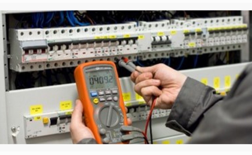 Miernik uniwersalny w dłoni elektryka, wyświetlający wartość pomiaru, na tle rozdzielnicy elektrycznej z bezpiecznikami i okablowaniem.