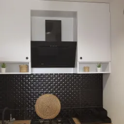 Nowoczesna kuchnia z czarnymi błyszczącymi płytkami 3D, białymi szafkami i czarnym okapem, uzupełniona o wiklinowe koszyczki i małe rośliny doniczkowe na półkach pod szafkami.