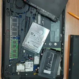 Otwarty laptop z widocznymi elementami wewnętrznymi: bateria, dysk twardy Toshiba, pamięć RAM. Obok leżą śrubokręty i fragment obudowy. Widoczne detale płyty głównej.