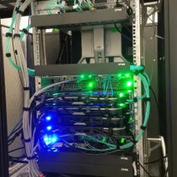 Widok z bliska na serwerownię w szafie rack: serwery Dell, macierz dyskowa i Data Domain Deduplikator Dell, przełączniki SAN i core, UPS Eaton, uporządkowane kable w kolorach szarym i turkusowym.