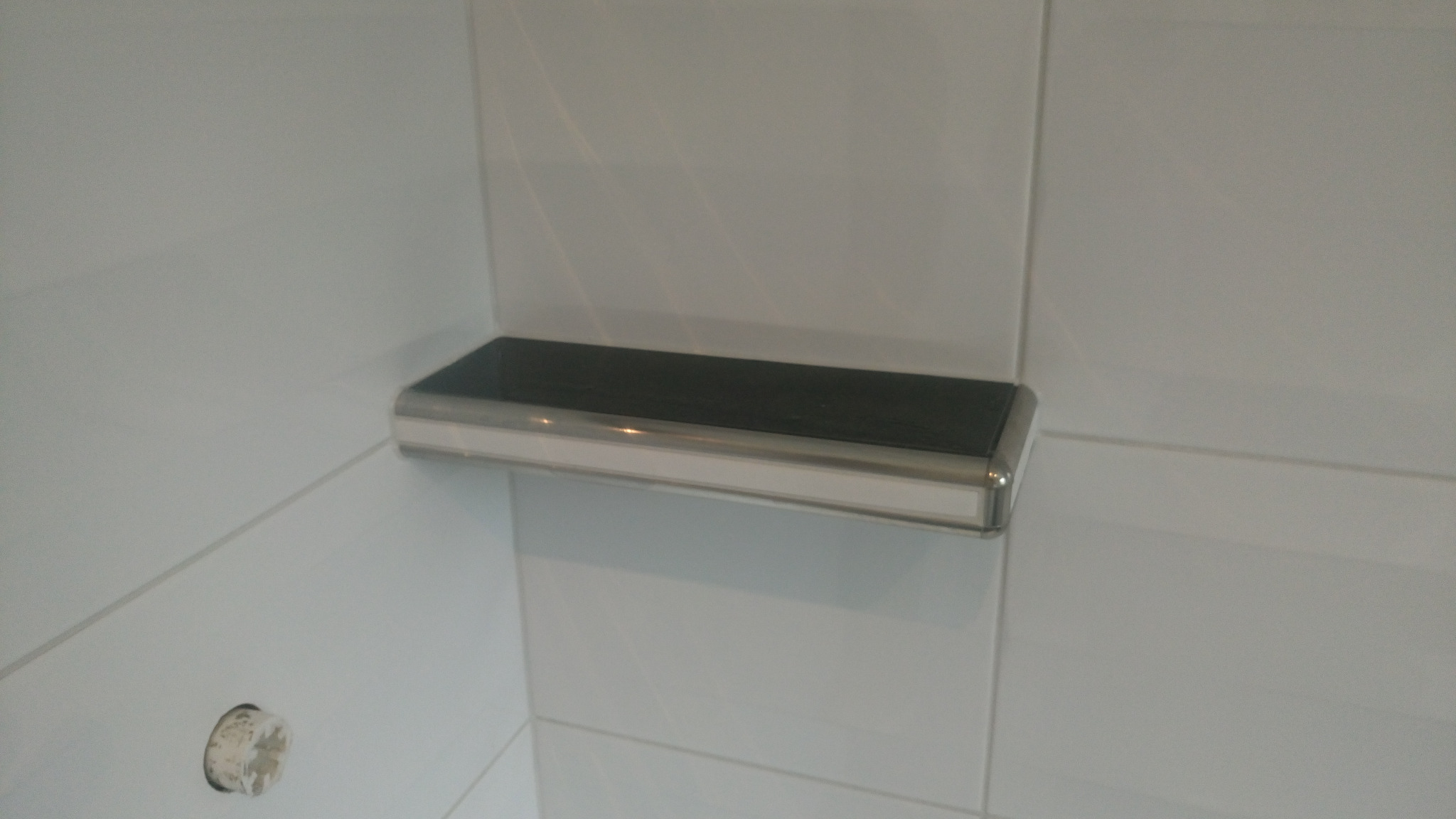 Półka łazienkowa z ciemnym blatem i chromowanym wykończeniem, zamontowana na białych płytkach ściennych, widoczny otwór po instalacji.