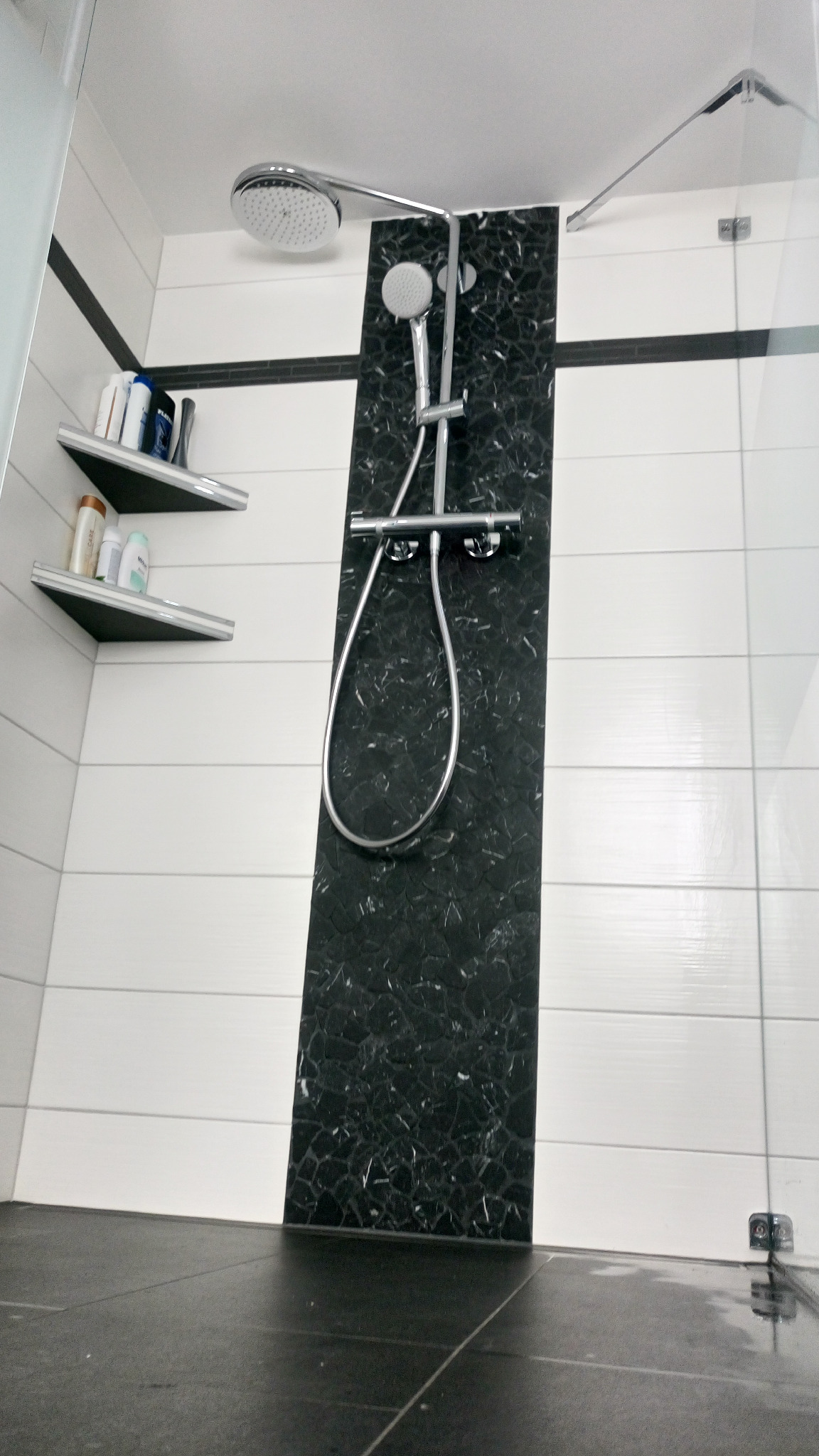 Nowoczesna kabina prysznicowa z panelem z czarnego marmuru, chromowaną armaturą i półkami z kosmetykami, widok z dołu.
