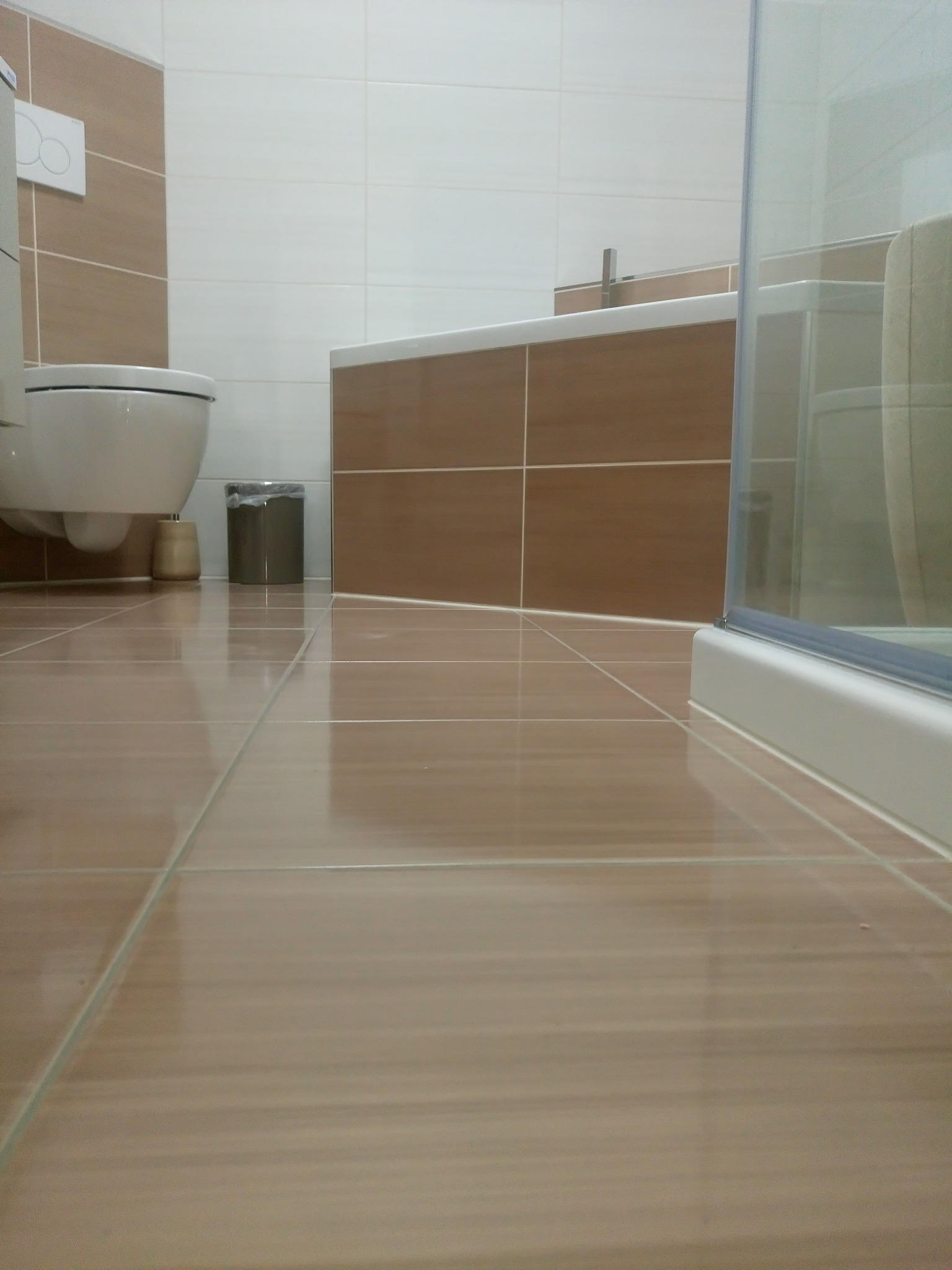 Beżowe płytki podłogowe w łazience, widok z niskiej perspektywy, toaleta wisząca i zabudowa wanny wykończone w brązach i bieli, minimalistyczny kosz na śmieci.