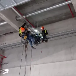 Dwóch pracowników w uprzężach, siedzących na metalowej platformie podwieszonej pod sufitem hali, instaluje koryta kablowe.