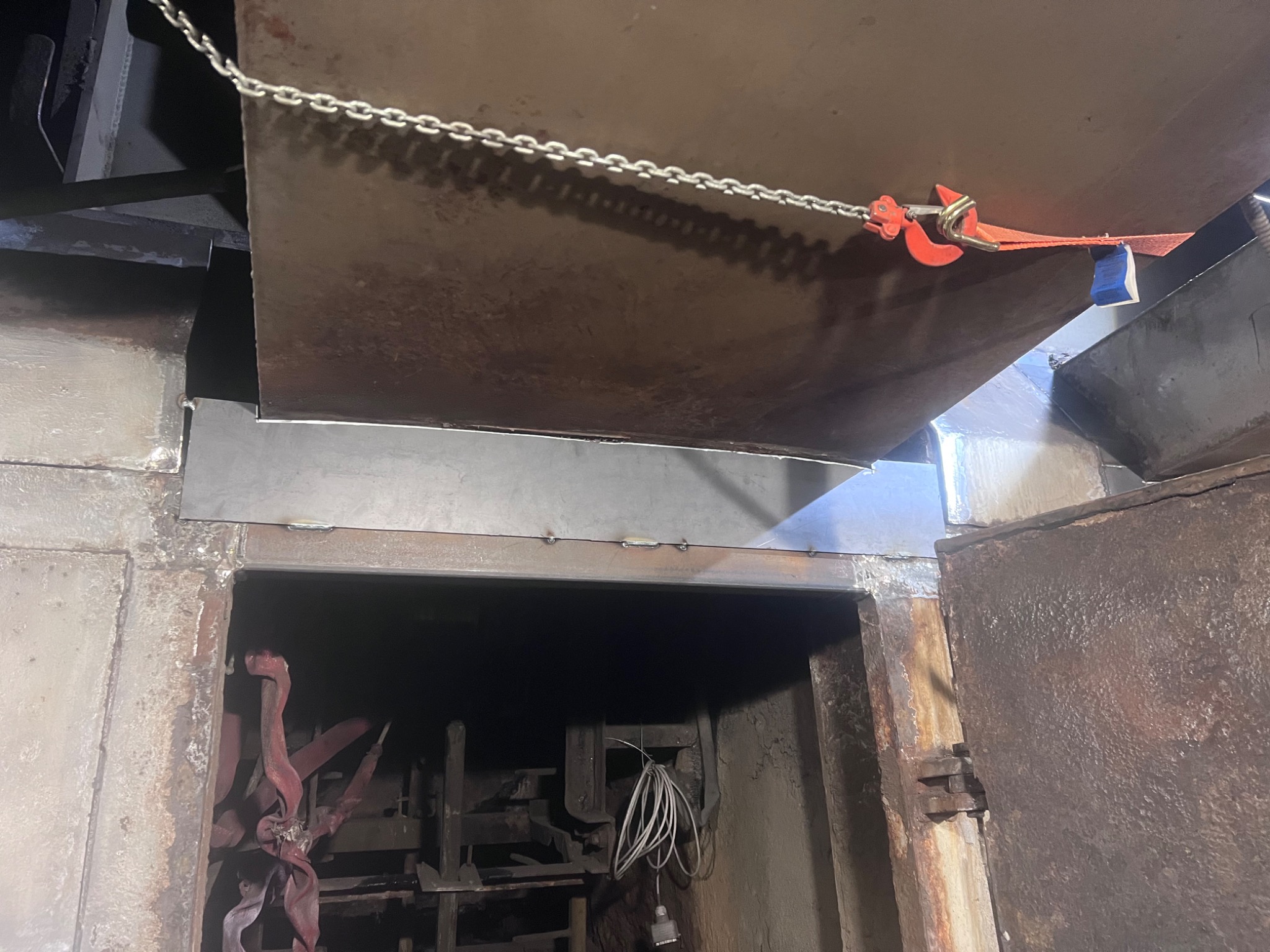 Metalowa płyta podwieszona na łańcuchu z czerwonym hakiem, częściowo zasłania wejście do metalowej komory z widocznymi elementami konstrukcyjnymi wewnątrz.