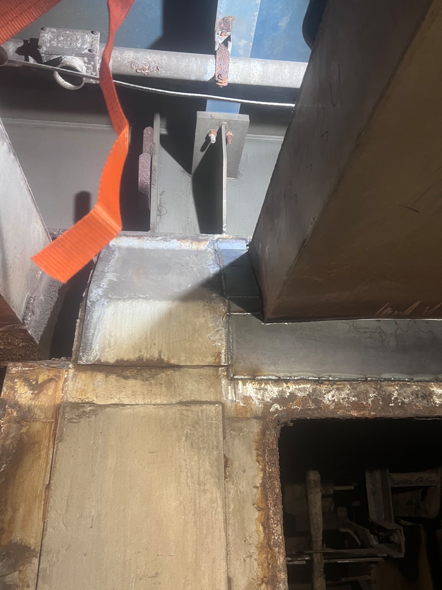 Spojenia spawane elementów konstrukcyjnych z metalu, widoczne ślady korozji i pomarańczowy pas transportowy.