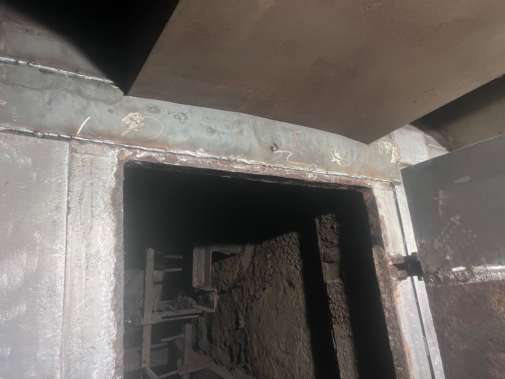 Metalowa konstrukcja z widocznymi śladami spawania, tworząca wejście do wnętrza z ziemistymi ścianami, oświetlona z góry.