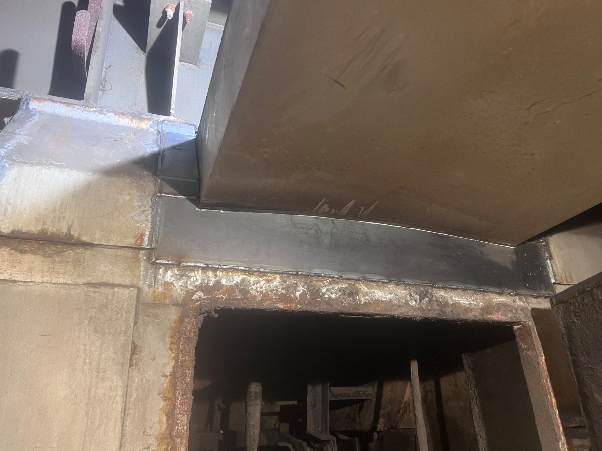 Metalowa konstrukcja po spawaniu, widoczne spoiny i ślady korozji na starym metalu. Nowo przyspawany element łączy się ze skorodowaną ramą, tworząc wzmocnienie nad otworem.