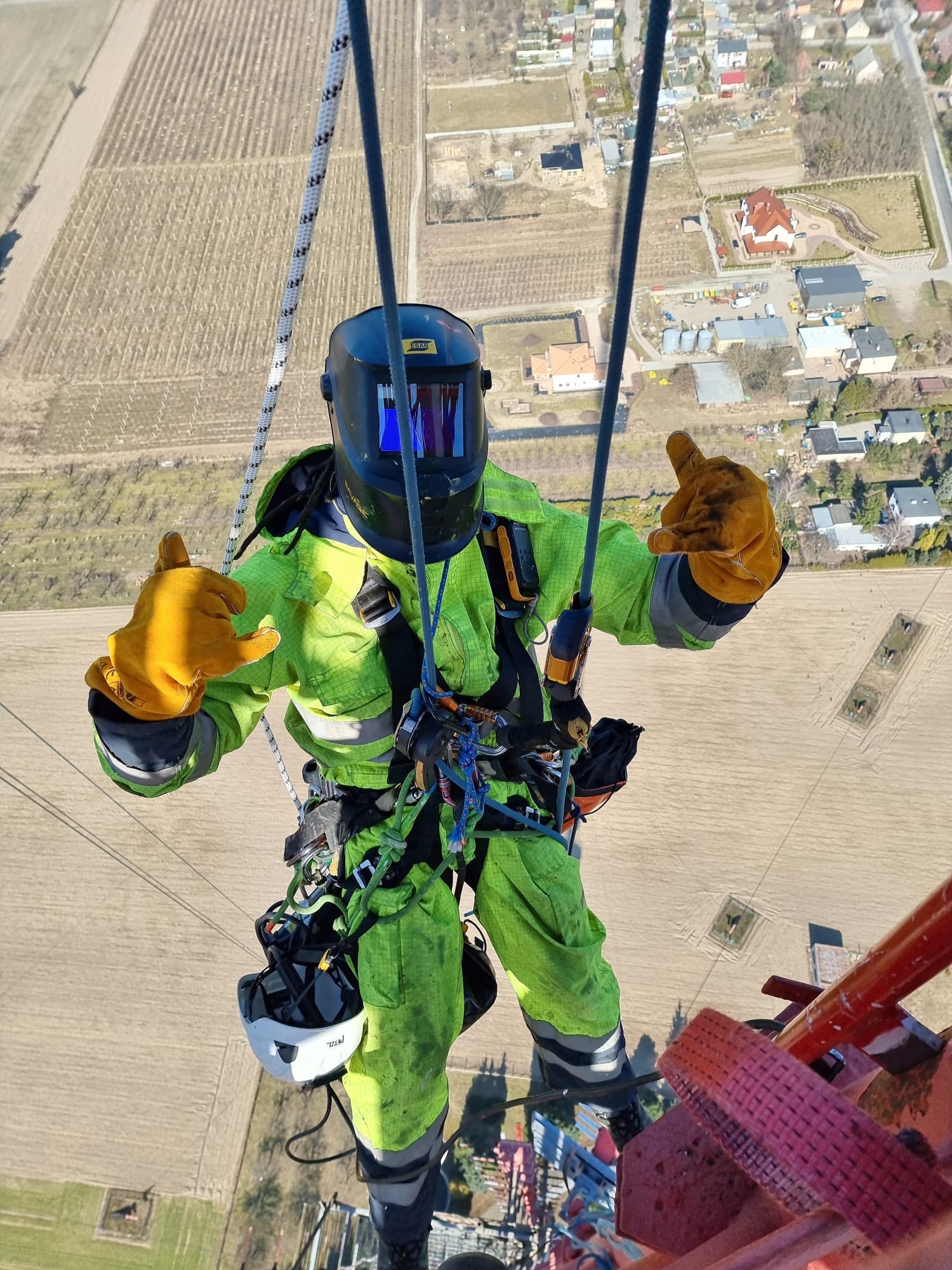 Pracownik w kombinezonie ochronnym i masce spawalniczej, zabezpieczony linami, wykonuje prace na wysokości, z widokiem na pola i zabudowania.