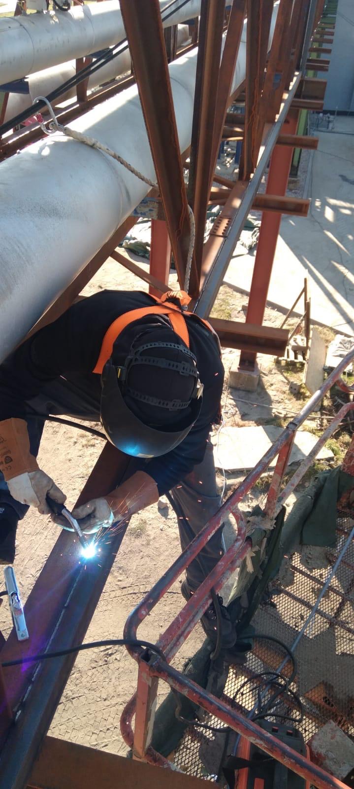 Pracownik w kasku spawalniczym i uprzęży zabezpieczającej spawa element konstrukcji stalowej na wysokości, iskry spawalnicze widoczne w jasnym świetle.