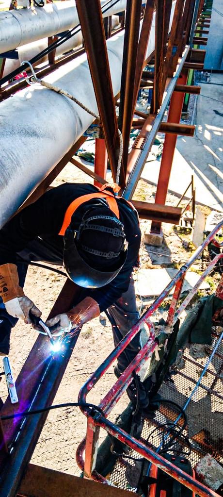 Pracownik w hełmie spawalniczym i rękawicach ochronnych spawa metalowy element konstrukcji na wysokości, widoczne iskry i spawarka, w tle rurociągi i elementy konstrukcyjne.