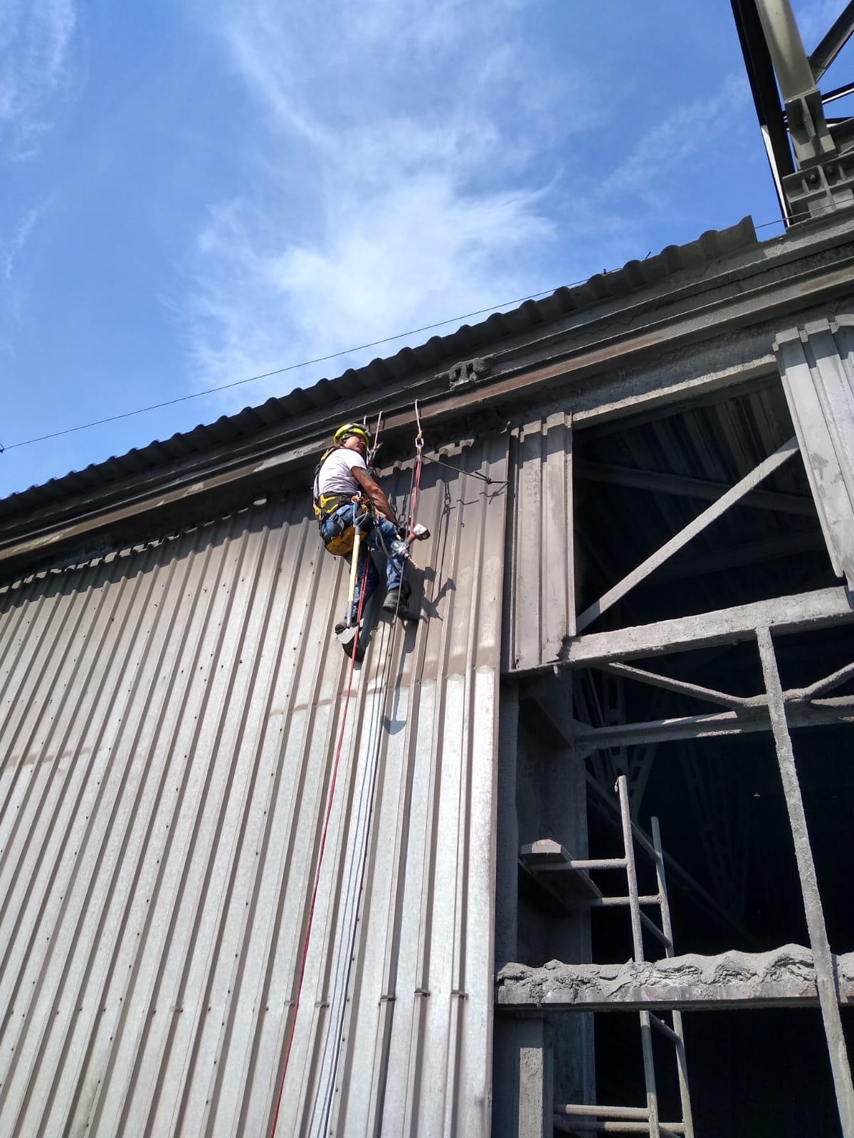 Pracownik w uprzęży, na linach, przy metalowej konstrukcji budynku przemysłowego, prawdopodobnie czyszczenie lub konserwacja elewacji.
