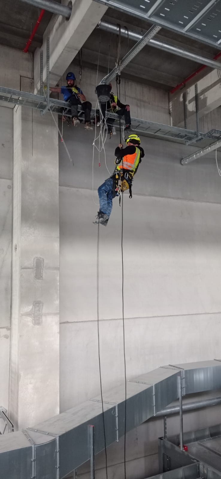 Pracownik w uprzęży wysokościowej, w kasku i kamizelce odblaskowej, opuszcza się na linie w hali przemysłowej, gdzie inni pracownicy siedzą na metalowej konstrukcji pod sufitem.