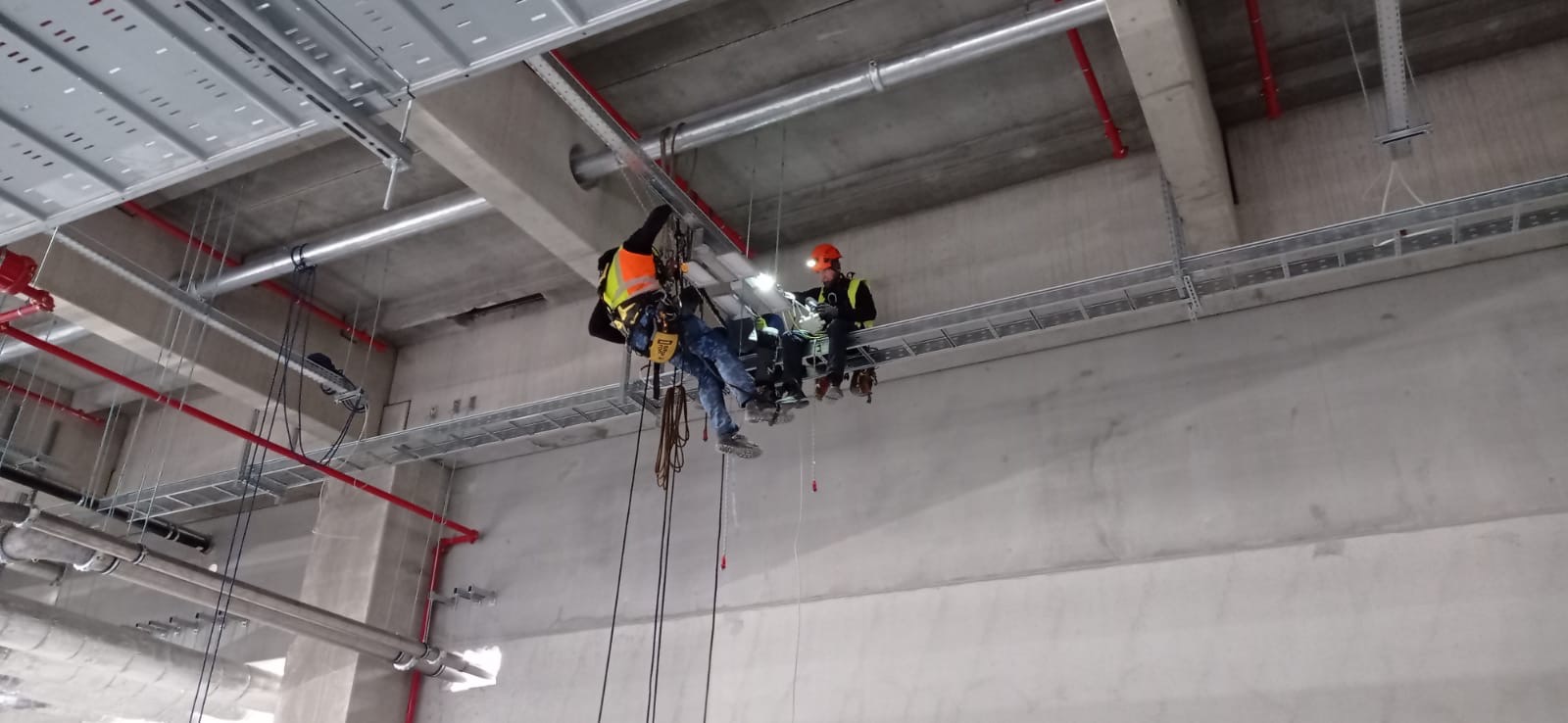 Dwóch pracowników w kaskach i kamizelkach odblaskowych, zawieszonych na linach, wykonuje prace na wysokości wewnątrz budynku przemysłowego, prawdopodobnie montaż instalacji pod sufitem.
