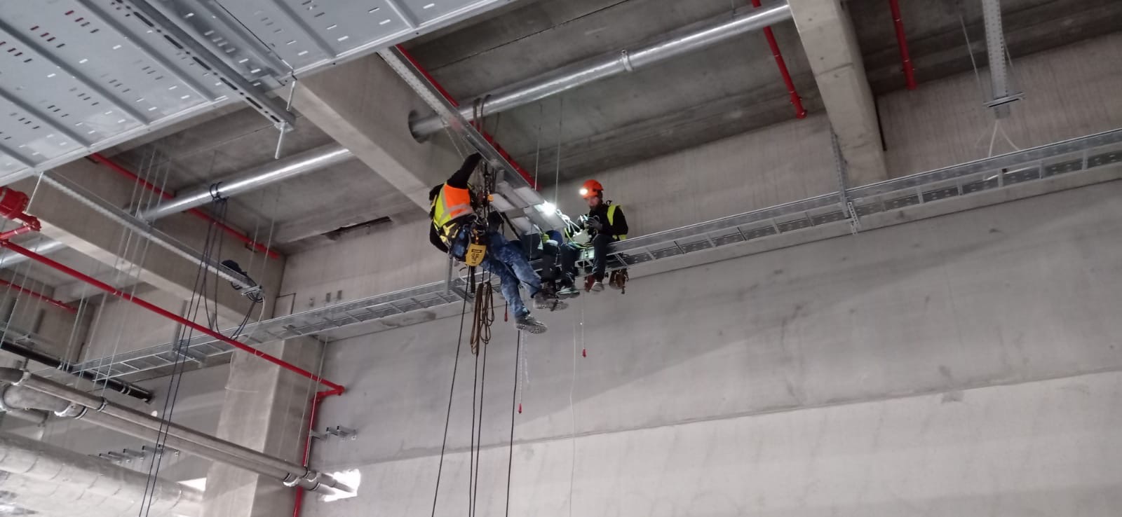 Dwóch pracowników w uprzężach, siedzących na metalowej platformie podwieszonej pod sufitem hali, instaluje koryta kablowe.