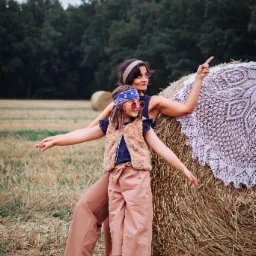 Matka z córką w stylizowanych strojach pozują na tle pola ze słomianymi balotami, dziewczynka w okularach i bandanie, kobieta wskazuje palcem w dal, na balocie leży biała, koronkowa narzuta.