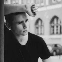 Czarno-białe zdjęcie młodego mężczyzny opierającego się o ścianę, z ręką na czole i zamyślonym spojrzeniem w bok, na rozmytym tle architektonicznym.