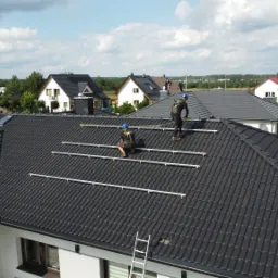 Montaż konstrukcji wsporczej pod panele słoneczne na dachu z ciemnej dachówki, dwóch instalatorów w kaskach i uprzężach zabezpieczających, drabina oparta o dach, w tle zabudowa jednorodzinna.