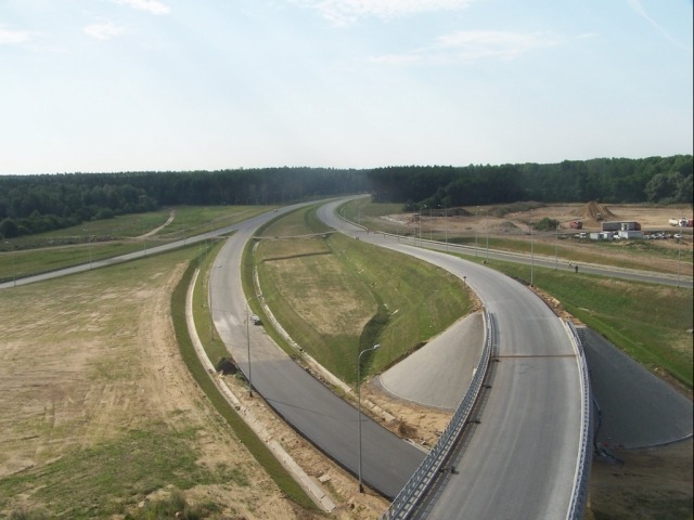 Zdjęcie z lotu ptaka przedstawiające fragment nowej drogi ekspresowej z wiaduktem, widoczne bariery energochłonne, oświetlenie drogowe i tereny zielone. W tle las i plac budowy z maszynami.