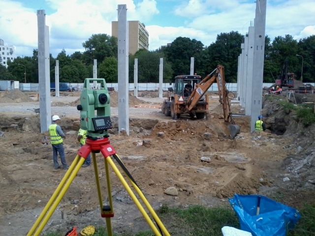 Zielony teodolit na żółtym statywie na tle placu budowy z betonowymi słupami i koparką