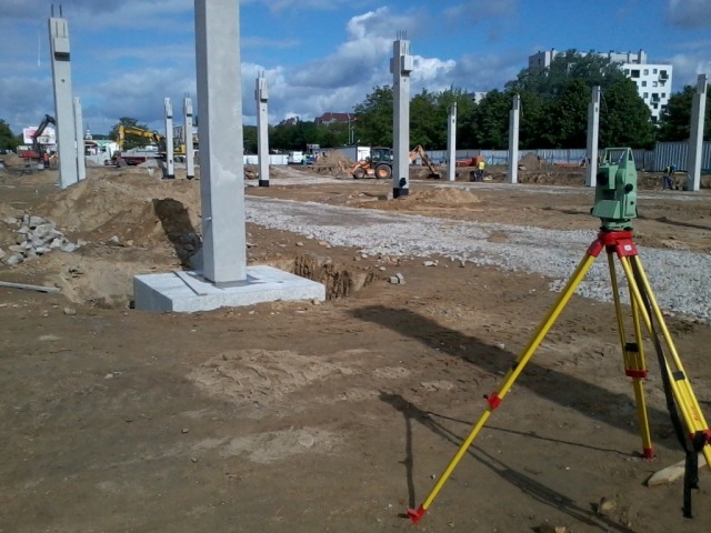 Prace ziemne pod budowę, widoczne betonowe słupy konstrukcyjne i niwelator optyczny na statywie z żółtymi nogami. W tle maszyny budowlane i budynki.