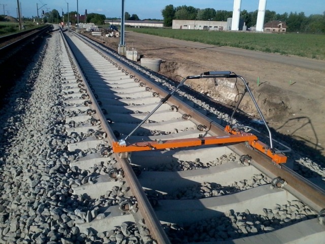 Pomarańczowe urządzenie pomiarowe na torach kolejowych, szyny pokryte tłuczniem, w tle widoczne budynki i zieleń.