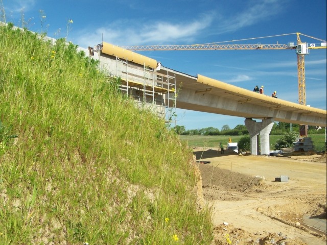 Budowa estakady w Pajęcznie: widok na betonową konstrukcję z żółtymi osłonami, rusztowania i żuraw na tle zielonego wzgórza i błękitnego nieba.