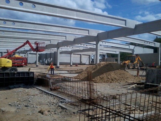 Budowa hali: widok na szkielet konstrukcyjny z betonu, zbrojenia, koparkę, podnośnik koszowy i pracownika z niwelatorem.