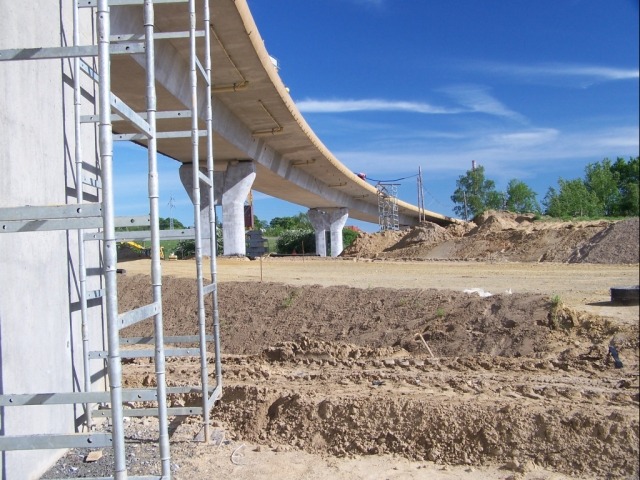 Budowa wiaduktu z widocznym rusztowaniem przy filarze i ziemnym nasypem w tle pod błękitnym niebem.