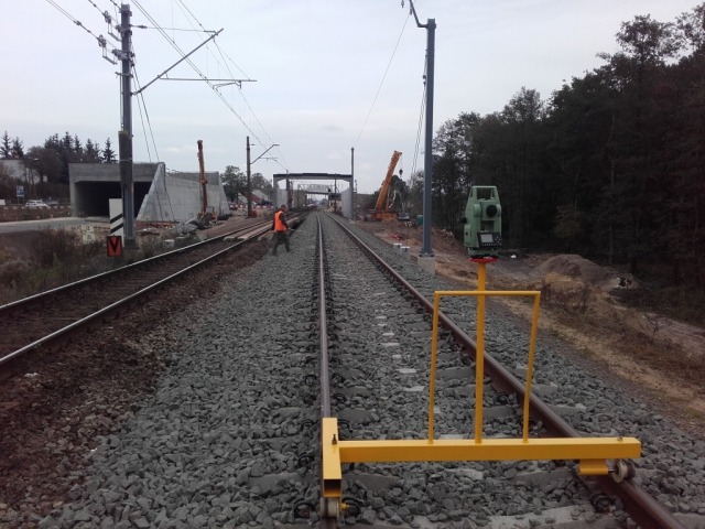 Prace pomiarowe na torach kolejowych z użyciem teodolitu na żółtym wózku, w tle widoczna budowa wiaduktu i pracownik w pomarańczowej kamizelce.